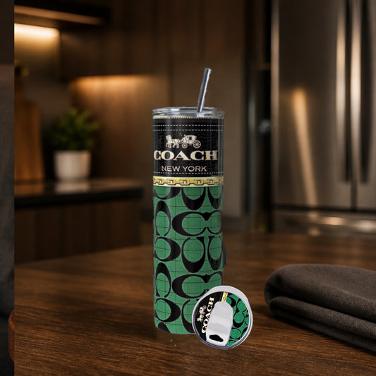 A tall insulated tumbler with a metal straw stands on a wooden countertop in a kitchen setting. The tumbler has a black upper section with gold and a green lower section covered in the signature pattern.