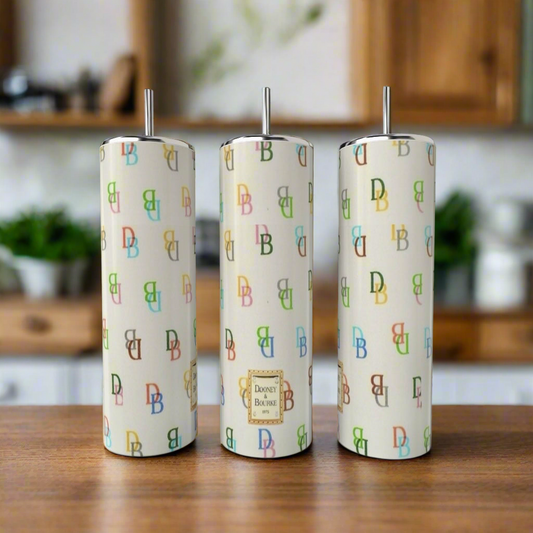 Three tall Kreative Kreationz D&B - Multicolor | Beige 20oz stainless steel tumblers with metal straws sit on a wooden counter, featuring a vibrant "DB" pattern and label—stylish, eco-friendly, and reusable against a blurred kitchen backdrop.