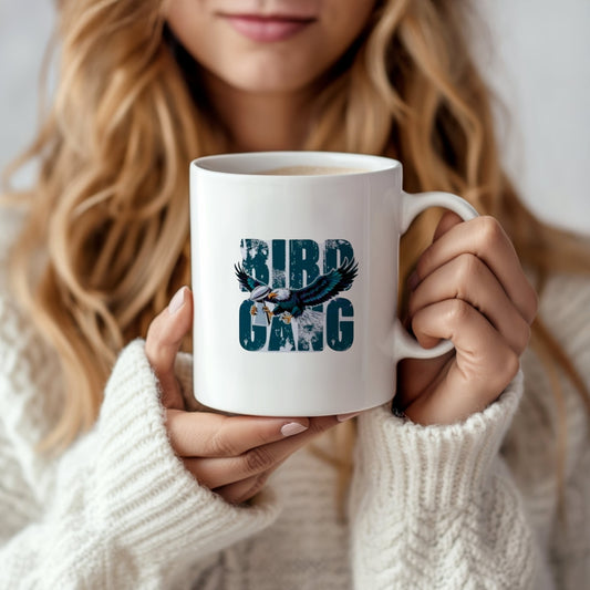A woman in a white sweater holds the 11oz "Bird Gang Ceramic Mug" by Kreative Kreationz, featuring a bird with outstretched wings—an ideal gift for Philadelphia Eagles fans. Only her hands and the lower part of her face are shown.