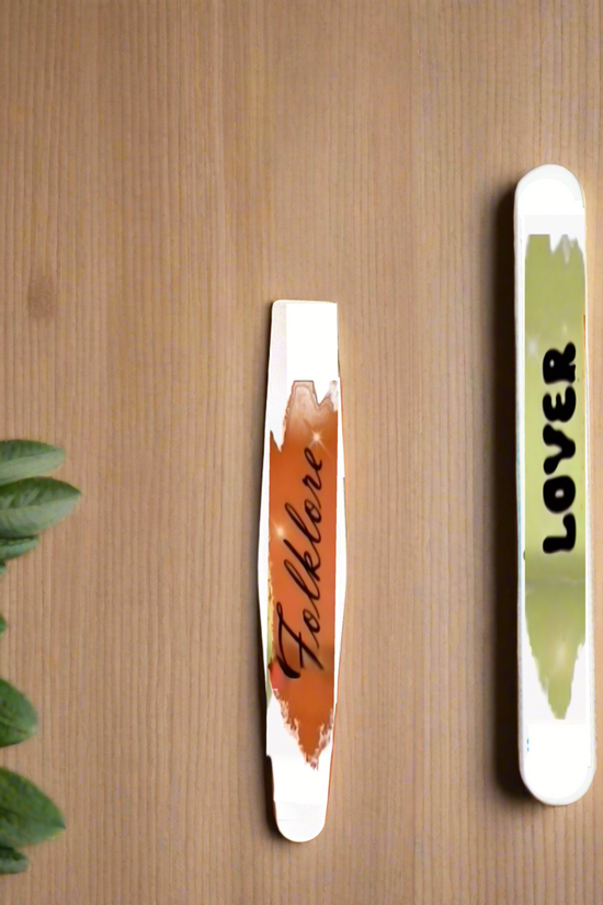 Two nail polish swatches, "Folklore" (brown) and "Lover" (light green), are shown on wood with green leaves—perfect Swifties nail art inspiration or to pair with your Kreative Kreationz Eras | 180 GRIT Stainless Steel Emery Board.