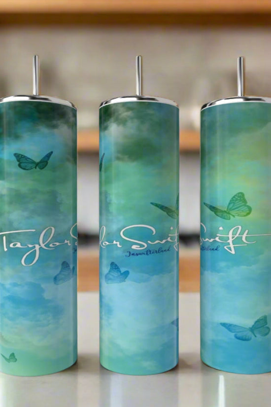 Three Debut Taylor Swift 20oz Tumblers by Kreative Kreationz with metal straws sit on a kitchen counter. The eco-friendly, stainless steel tumblers feature a blue-green gradient, butterfly designs, and white “Taylor Swift” text.