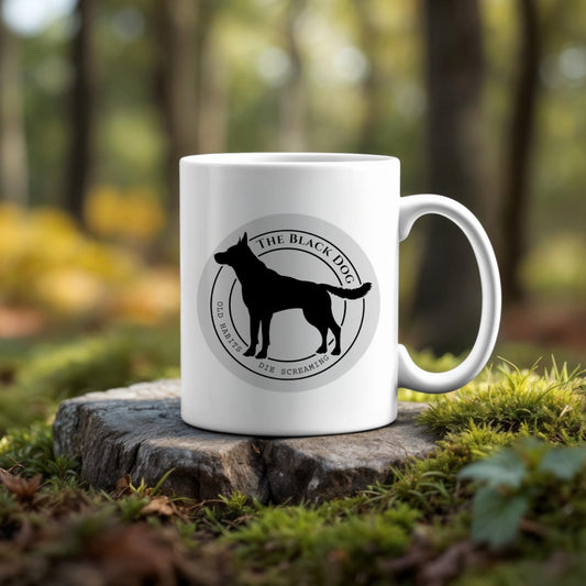 A TTPD - Taylor Swift Mug by Kreative Kreationz featuring a black dog silhouette and "The Black Dog – Die Happy, Die Screaming" sits on a tree stump in a forest, perfect for unique Swiftie collections.